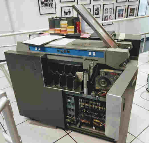 The IBM 1402 Card Reader/Punch is a large piece of equipment. It has a metal chute to load cards and puts cards into various hoppers in the middle. It has a few buttons and lights for control. A door on the front is open, revealing the circuitry inside.
