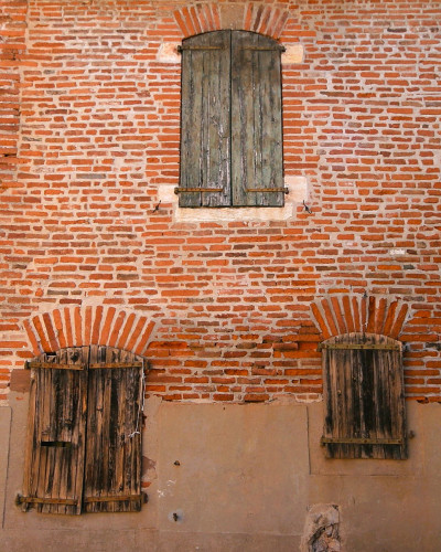 Three shuttered windows in a redbrick wall - one above and two below. The windows are all different sizes and the shutters are unpainted. Nothing seems to be in a very good state of repair.