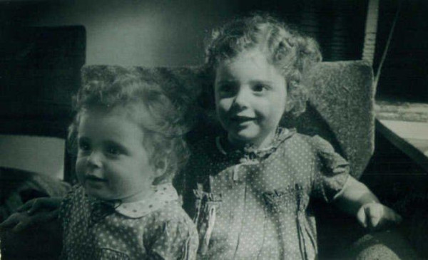 Vintage black-and-white photo of young sister closely together, both dressed in dotted dresses and with curly hair, looking towards the camera.