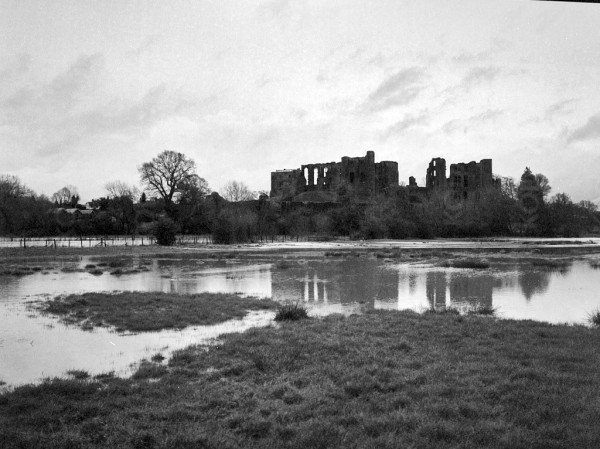Black and white photo showing a ruined castle reflected in flood waters, with a ragged sky above. There is little detail in the scene from this grey day.