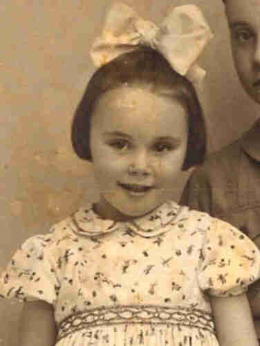 A young girl visible from her waist up. She is wearing a dress with short sleeve with some toy patterns. She has dark hair reaching her ears and a huge white ribbon in he hair. 