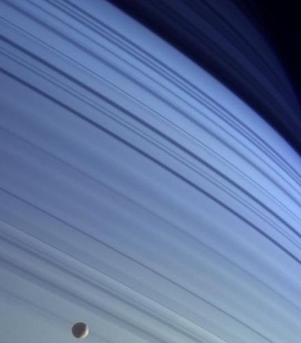 The shadows of Saturn's rings on the hazy, blue surface of the gas giant dominate the entirety of this picture, spanning from almost white in the bottom left corner to the black of night in the top right corner. Tiny moon Mimas is seen in the bottom left against the Saturnian backdrop.