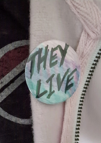 "They Live" in the 80s movie font, written on a button with a pink-to-blue gradient background, pinned to a light pink hoodie, over a black Pink Floyd t-shirt.