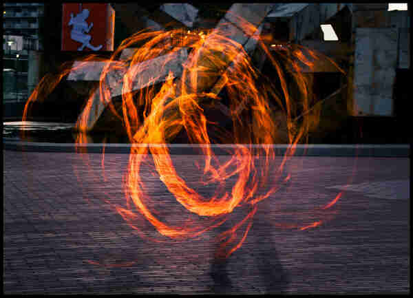 A swirling, tangled ball of fire surrounds the shadowy wisp of a person on the Embarcadero Center plaza. Although.taken at night, the orange flames do little to illuminate the brick plaza or the concrete fountain in background.