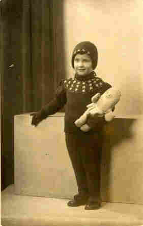 A girl of several years standing in the atelier. She is dressed in a hat, jumper, gloves and long trousers. Stars can be seen on the jumper. Under her right arm she holds a large toy - an oval-shaped doll. She rests one hand on the cuboid that forms the background.
