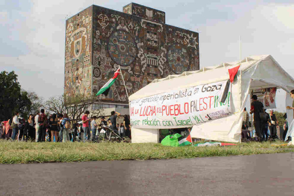 People gather near a white tent with a pro-Palestine banner and Palestinian flags on the campus of UNAM. In the background, one can see the mosaic-covered facade of the Central Library.