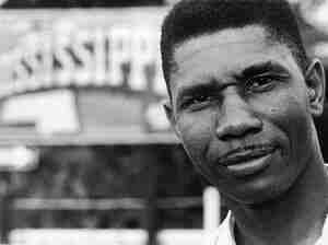 Black and white photo of Medgar Evers, from the neck up, looking straight at the camera. There is an out of focus sign in the background that says “Mississippi.”