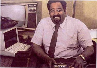 Low-quality photo of a smiling Jerry Lawson from the early 1980's, sitting next to a computer and television set and holding the game system he invented. Lawson is a black man in his early 40's with a goatee, wearing a white short-sleeve dress shirt and black tie.