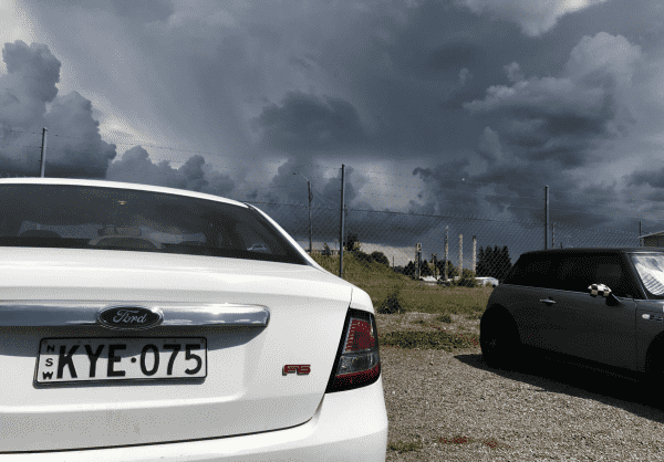 A photo of a couple of cars close-up in sun, with an absolutely epic set of dark, rising, ominous and near blue clouds over towards the horizon.