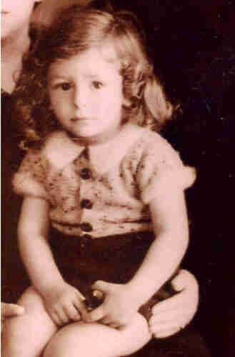 A young boy with long cury hair sitting on her mother laps. He has short sleeves and short trouser. He is holding something in his hands.