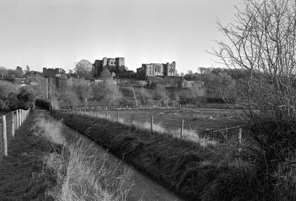 A ruined castle dominates the horizon of this black and white photo, with three large buildins evident. In the  foreground a sunken lane goes from bottom right to centre left, bordered by fence posts above the bank on either side. A bare thorn bush grows on the right side.