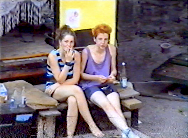 Zwei junge Frauen sitzen vor einem zum Wohnen ausgebauten Bauwagen, aus der Ferne von schräg oben fotografiert. Beide tragen kurze Hosen oder Röcke und Trägerhemden. Eine zieht an einer Zigarette und blickt amüsiert in die Kamera.