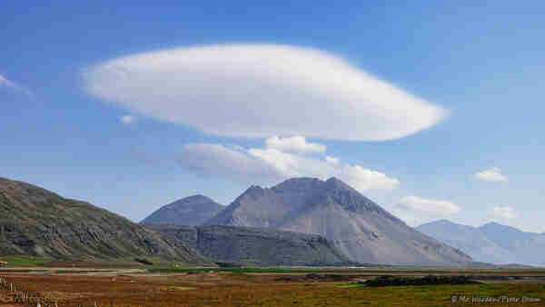 A photo of a summer scene with blue sky and fluffy clouds. The foreground is brown vegetation but some green can be seen at ground level in the distance. On the horizon is a rugged bare mountain with grey flanks and a caldera-shaped saddle at the top. Hanging above the summit is a large disk-shaped lentoid cloud, pure white above and slightly greyish below. Its surface is smooth and almost featureless. The scene is lit from the right.