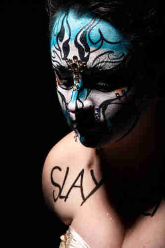 Coles up portrait  of a drag queer wearing blue and white make up.
She has the word "Slay" written on her shoulder.