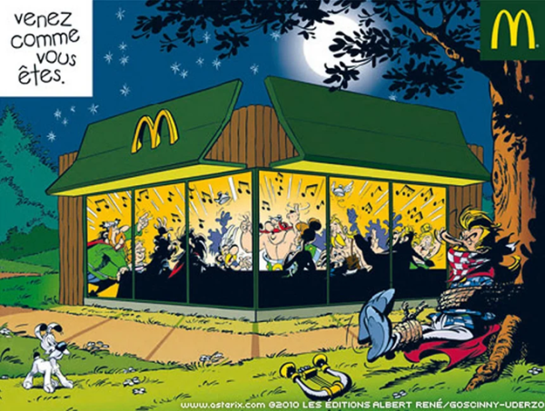 Die Gallier beim Festbankett, jedoch nicht an einem Lagerfeuer mit Wildschwein, sondern in einem erleuchteten McDonald's Restaurant. Troubadix sitzt gefesselt draußen an einem Baum.