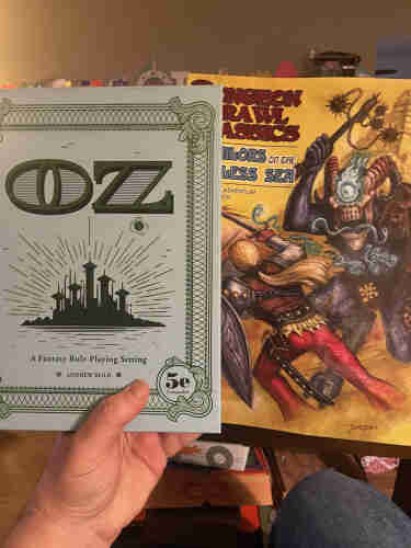 Two role-playing game books held by a person against a background of a room. The left book's cover says "OZ - A Fantasy Role-Playing Setting" and indicates it is compatible with 5e, while the right book features colorful artwork.