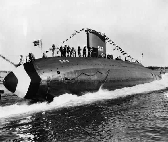 A black and white photo courtesy of the U.S. Navy that shows the launch of the USS ALBACORE in 1953. Celebratory flags fly from the large submarine as crew members stands on the top of the vessel during her first time at sea.