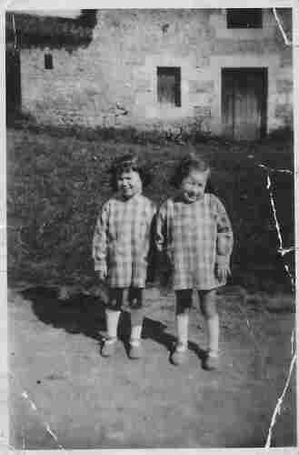 A photo of two young tween girls standing outside. There is a lawn and house behind them. They have checked dresses and long socks. They are looking at the camera.
