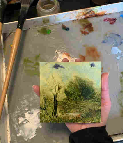 Photo of my hand holding a 4”x4” landscape oil painting over a paint palette. The painting is a color study, experimenting with different greens, blues, and reds to create a rough landscape scene. Puddles of oil paint are visible on the paint palette, paint brushes rest on the left edge of the palette box. A jar of linseed oil is visible at the top of the photo.