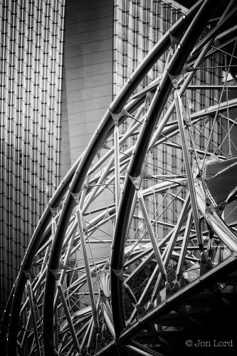A Black And White Photo In Portrait Format. In The Foreground And To The Top Right - Descending Down To The Bottom Left Is A Lattice Like Structure. On The Outside Are Curved Non-Reflective Tubes Surrounding The Structure In A Spiral Shape From The Base On One Side To The Base On The Opposite Side. Within The Spirals Are Shiny, Perhaps Stainless Steel, Rods That Support The Base. Behind Is The Front Elevation Of A High-Rise Building. It Is Glass Fronted With Vertical Strips Stretching From The Bottom To The Top. The Lattice Structure Is The Double Helix Bridge That Crosses Marina Bay And The Building Is The Marina Bay Sands Hotel. There Is No One Visible In The Image. 

Marina Bay, Singapore, 2013