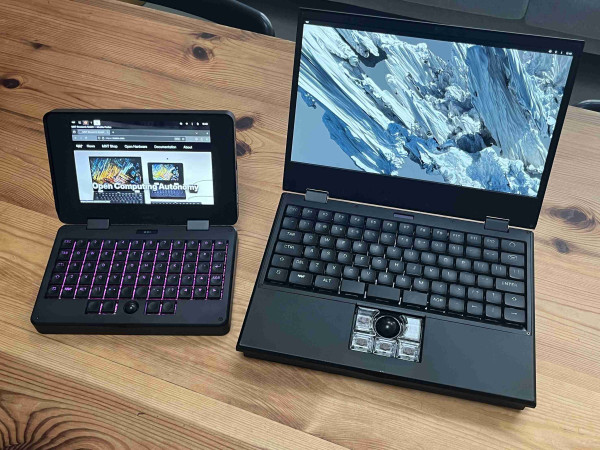 a cute 7 inch laptop with colorful keyboard backlight that has a bit of an industrial vibe, next to a bigger one that has more of an industrial vibe and a snazzy trackball. both rest on a wooden table surface