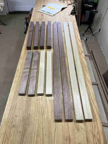 Assorted wooden pieces arranged on a workbench in a workshop with an open notebook. The wood is dark walnut and light poplar. 