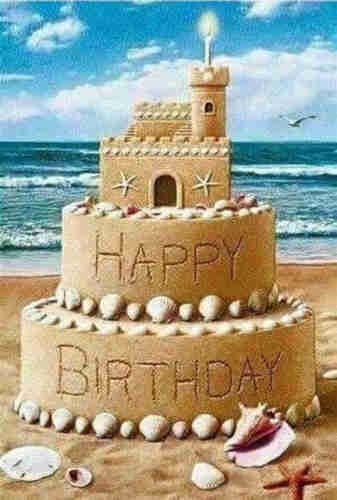 A birthday cake sand sculpture with sea shells around the bottom of each of 2 tiers and a small sand castle on top. The ocean is in the background.
