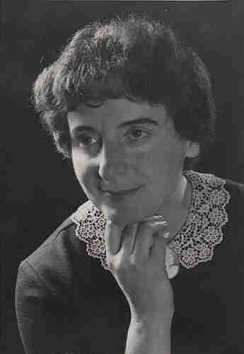 Black and white photograph of a mature woman. She is wearing a blouse with an embroidered collar. She supports her head with her right hand - her chin rests on her palm. She is smiling slightly. She has medium-length dark hair.