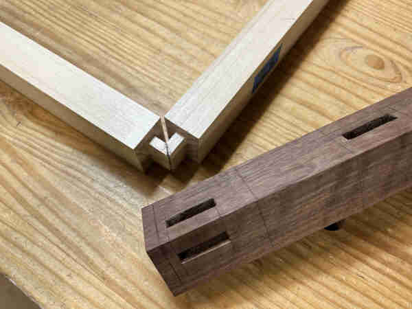 Sitting on top of a woodworking bench, two long pieces of light colored wood meeting at a 90 degree angle from their ends. There is a tenon on each end with a miter and the tenons fit together.