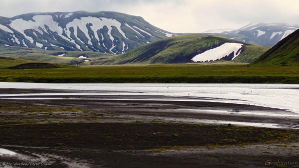 A photo of a landscape, with the muddy shore of a lake in front of the viewer. Small birds are flying above the water surface. The opposite shore is green with vegetation and, where the substrate shows through, dark bluish grey. Some sheep can be seen grazing in the meadow. Large patches of snow and ice lie between the ridges of the mountain faces beyond, creating an odd patchwork of scalloped shapes again the dark rock and soil beneath and the green vegetation above. It's strangely lovely, and quite otherworldly.