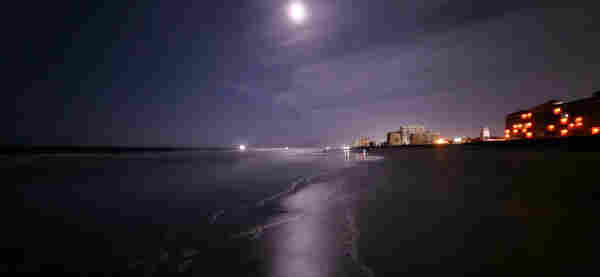 Late night looking out over a low tide beach. A slow, but steady flow of light waves wash ashore leaving damp sand and white bubbly trails behind. High in the dark night sky the moon glows bright, shining a long reflection down upon the coastline surf. Surrounding the moon is a light layer of thin, smoke like clouds. The low tide, compact sand leads up to fluffy, loose sand along the shore before a long row of coastal buildings illuminated with night lighting.