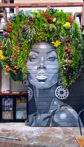 Streetartwall. The mural of an African-American woman was created with paint and flowers on the red brick wall of a café terrace. The beautiful black woman is painted in black, white and gray. She has fine facial features, wears large round earrings and looks directly at the viewer. Her Afro hairstyle, on the other hand, is a colorful sea of flowers made up of arranged green tendrils and leaves and petals in red, yellow and purple. A beautifully designed portrait of a woman (in the photo there is a small drinks cabinet next to the mural).