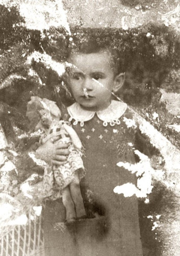 A vintage, black-and-white photo showing a young girl in a dress, holding a doll. The photo is aged and damaged, with visible scratches and white spots.
