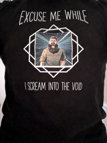 A black T-shirt with a cartoon picture of a man screaming with the caption, "excuse me while I scream into the void"