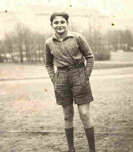 A photo of a teenage boy standing outside. He is wearing shorts. He keeps his hands in his pockets. Behind him, grass and some trees.