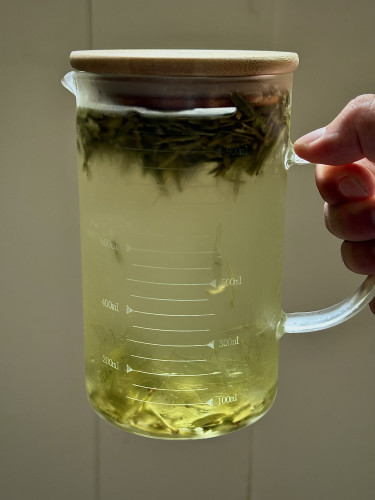 My hand holding a 1 liter glass pitcher with bamboo lid filled with cold brewed green tea. The leaves are mostly floating at the top but some have fallen to the bottom. 