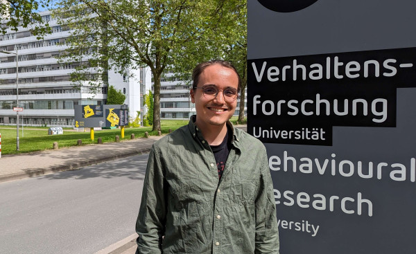 Young white man standing in front of the buildings of Bielefeld University.