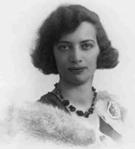 Portrait photograph of a young woman. She has long wavy hair with a wide fringe. She has a wide lips. There are beads around her neck.  She wears a dark blouse and a fur shawl.