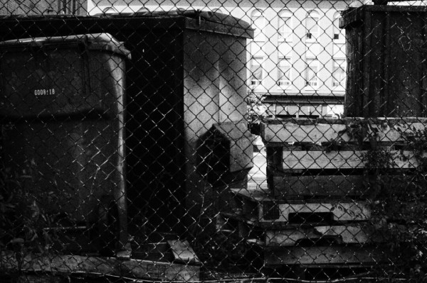 Garbage cans and assorted scraps sit behind a chain link fence in this moody black and white image.