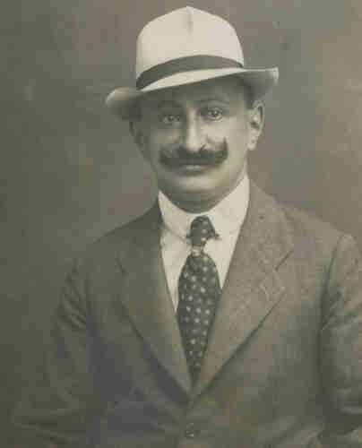 Portrait photograph of a mature man. He is wearing a jacket, shirt and tie with dotted patterns. He is wearing a light-coloured hat with a dark band. His moustache is curled upwards.