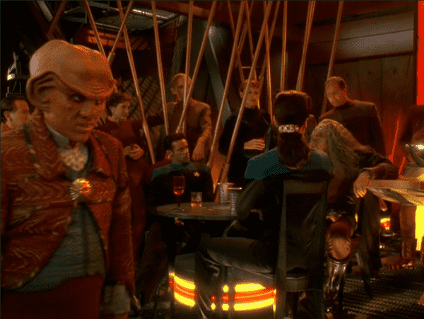 an image from the show, an old klingon regaling the crew and patrons of quark's bar, quark is on the left side with a sour expression on his face 