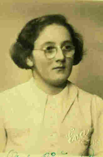 An ID portrait photo of a mature woman. She has long wavy hair covering her ears. She is wearing round glasses. Her shirt is buttoned to her neck. 