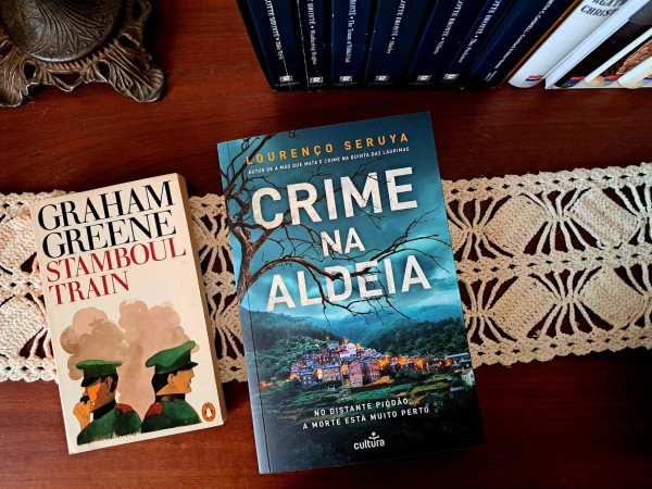Two books over a table, Stamboul Train by Graham Greene in a penguin edition and a Portuguese book whose title can be translated as Crime in the Village