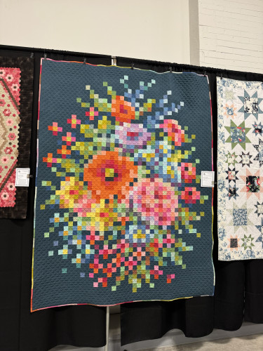 Quilt on display featuring a colorful, pixelated floral pattern against a dark background, hung between two other quilts.