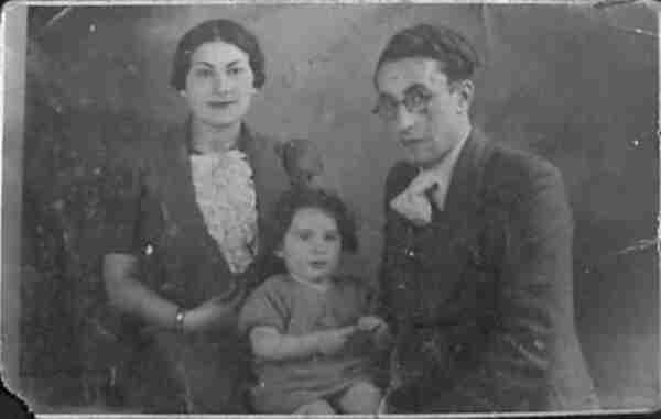 Family photo. From left: woman in jacket, blouse with frilly embellishments. She has dark hair combed to the side and pearl earrings; A little girl sitting and a man wearing a jacket and round-rimmed glasses.