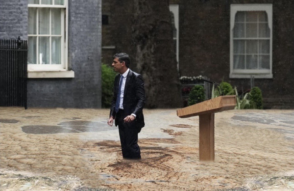 A soaking wet suited Rish! standing in knee-deep sewage floodwater beside a partly submerged wooden podium outside Number 10. It is raining.