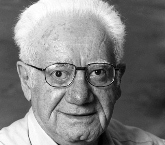 Portrait of an elderly individual with white hair and glasses, smiling slightly in a close-up black and white photograph.