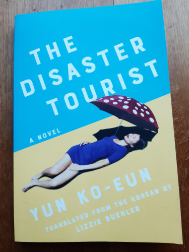 Book cover: top half blue, bottom half yellow. On the yellow half, a woman in a purple dress lying with eyes closed, her head in the shade of a red parasol with white spots. Title, author and translator in white. 