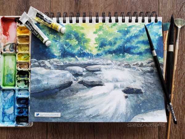 A photo of a watercolor painting in a spiral sketchbook of a nature scene with a vibrant forest in the top third with a stream running through rocks taking up the bulk of the painting. Paint and brushes lay next to the sketchbook.