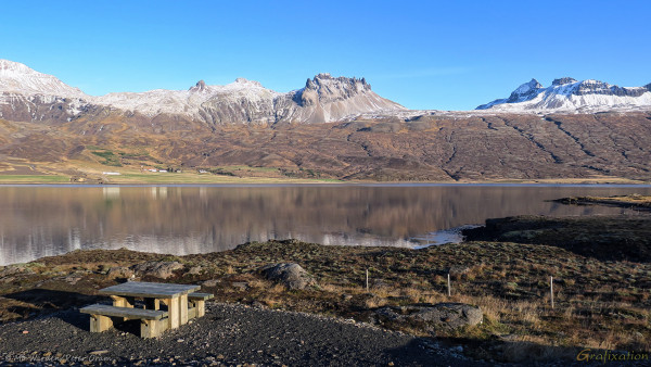 A photo of a landscape, with a clear blue sky overhead and sunlight is from the right. In the distance is a line of craggy mountains with a dusting of snow at the peaks. At their foot is a collection of farm buildings near the shores of a still fjord, which is reflecting the mountains. The foreground is a rugged, rocky waterline, with large stones and a fence outlining a gravel-covered area. On this level surface is an inviting wooden picnic bench.